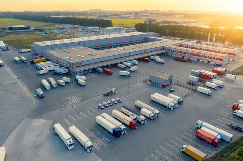 Logistics park with warehouse, loading hub and many semi trucks with cargo trailers standing at the ramps for load/unload goods at sunset.